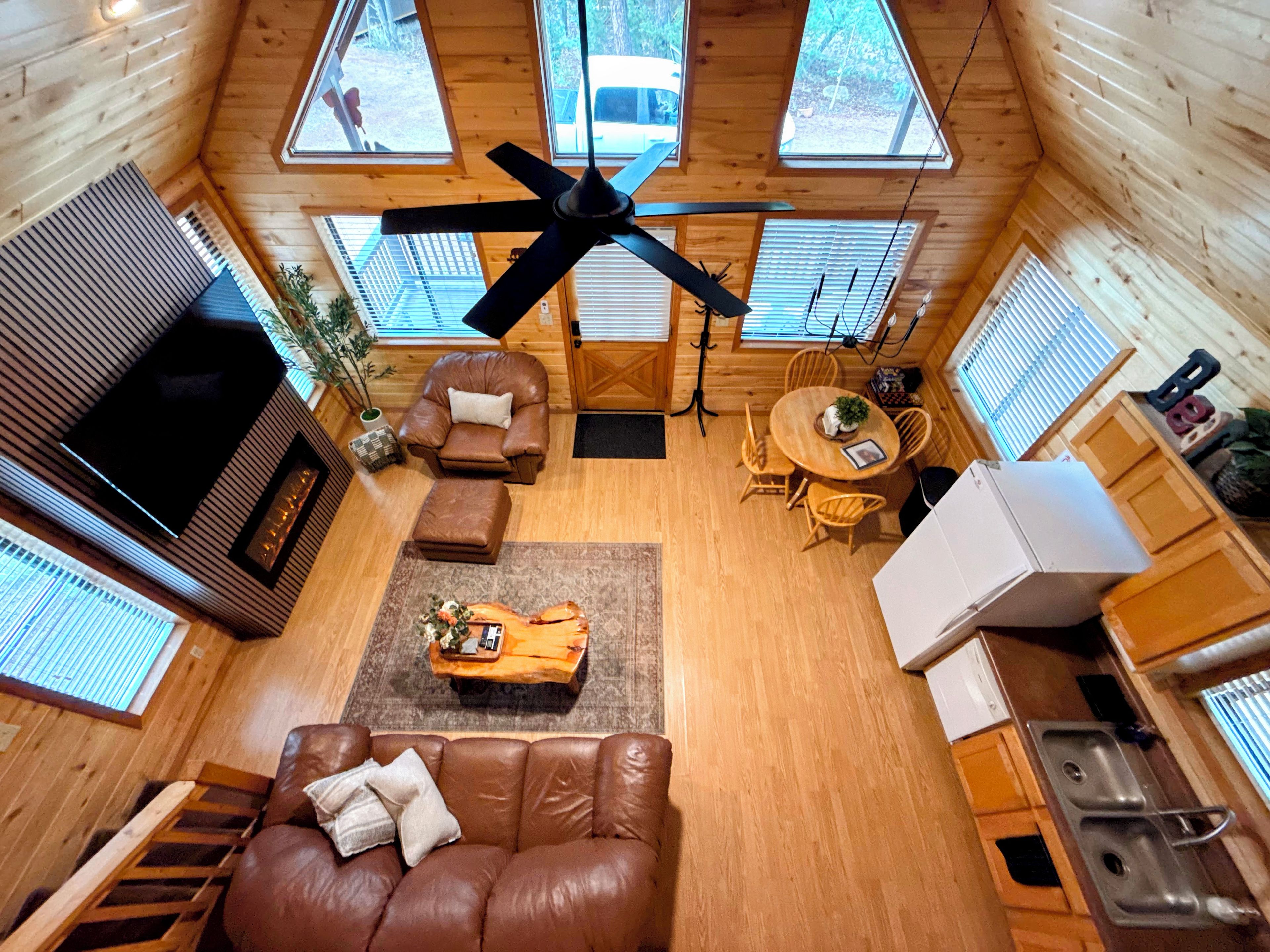 A cabin interior with a ceiling fan, wooden walls, a sofa, a dining table, and a kitchen area.