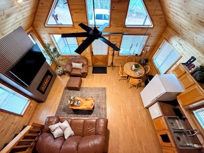 A cabin interior with a ceiling fan, wooden walls, a sofa, a dining table, and a kitchen area.