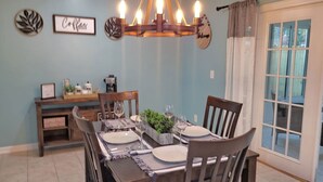 Uma área de jantar com uma mesa de madeira posta para quatro pessoas, rodeada por cadeiras. Há decorações nas paredes, um candelabro e uma vista pela janela.
