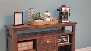 Uma estação de café de madeira com cafeteira, cesta, planta e um letreiro que diz “CAFÉ PRIMEIRO, PESSOAS DEPOIS”.