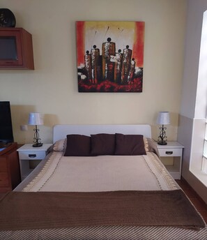 A bedroom with a bed, two bedside tables, a lamp on each table, a painting on the wall, and a window. - Iter Away Dunes (La Oliva)