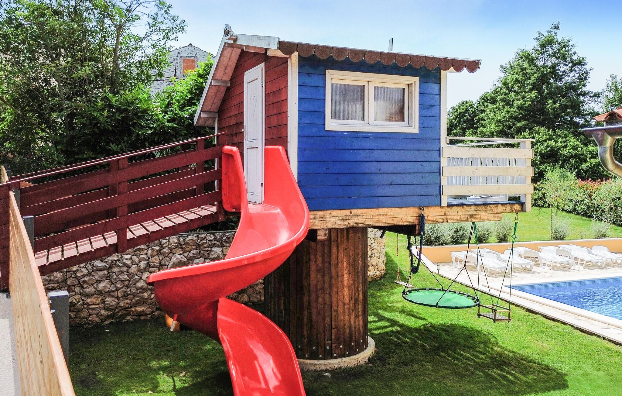 A wooden playhouse with a red slide, a stone wall, and a pool area.