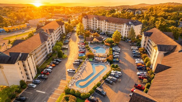 A multi-story hotel with a central swimming pool, parking lot, and surrounding greenery.
