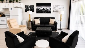 Una sala de estar moderna con un sofá negro, dos sillones negros, un sillón beige y una mesa de centro blanca, con una cocina al fondo.