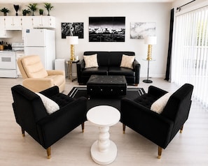 A modern living room with a black sofa, two black armchairs, a beige armchair, a white coffee table, and a kitchen in the background.