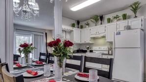Un comedor con una mesa dispuesta para cuatro personas, con servilletas rojas, copas de vidrio transparente y un florero con rosas rojas. Se ven armarios blancos y un refrigerador blanco al fondo.