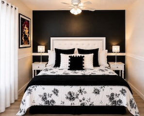 A bedroom with a white headboard, black and white floral bedding, two bedside lamps, and a ceiling fan.