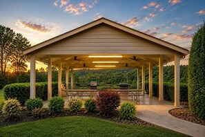 A covered outdoor area with picnic tables, a grill, and a view of a forested hillside.