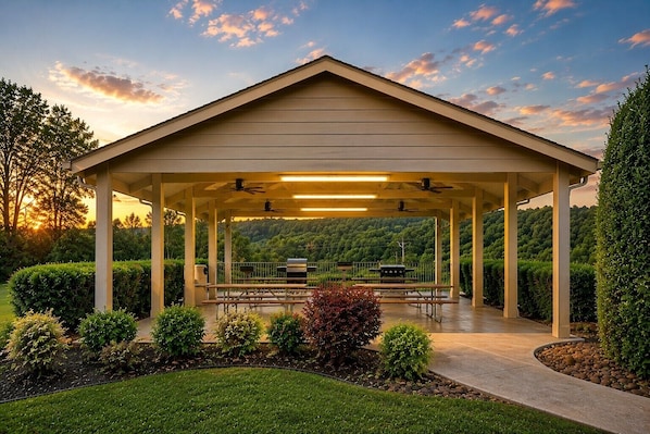A covered outdoor area with picnic tables, a grill, and a view of a forested hillside.