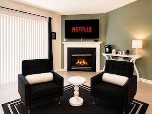 A living room with two black armchairs, a white coffee table, a fireplace, and a TV mounted above it displaying 'NETFLIX'.