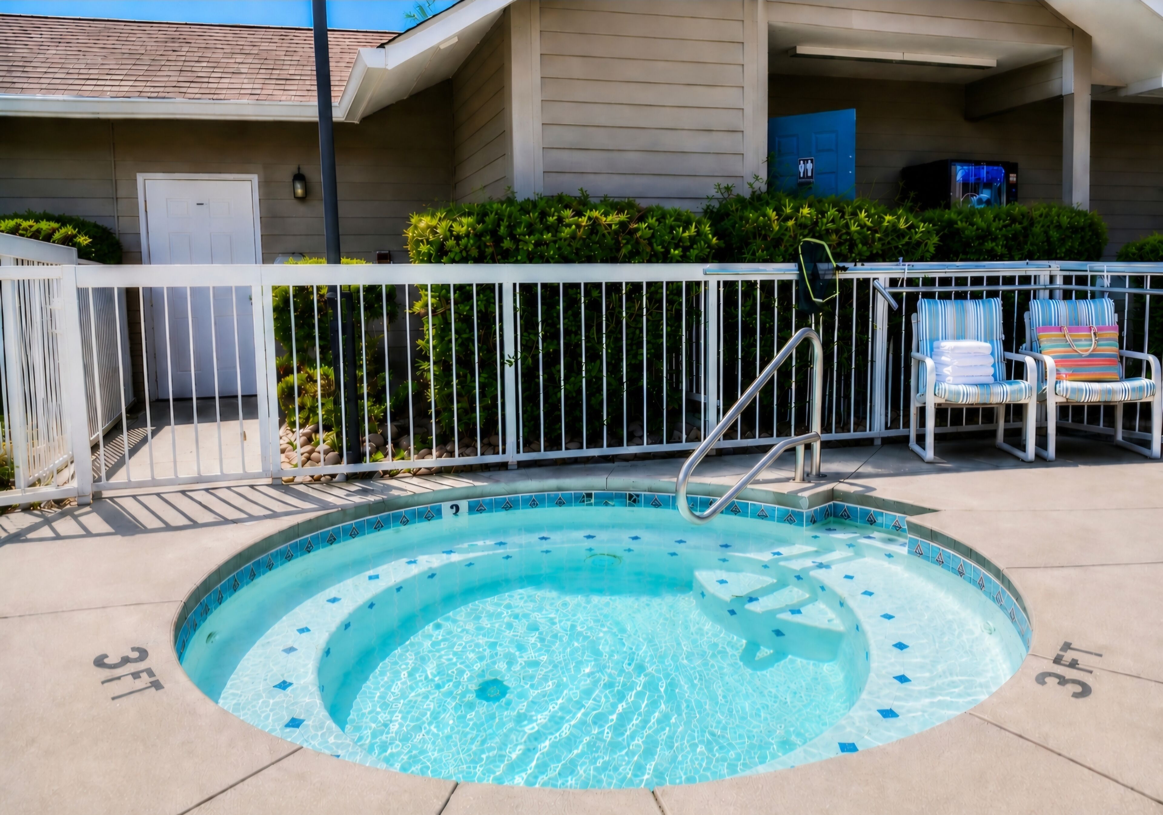 Une piscine de 90 cm de profondeur, entourée d’une clôture en métal, avec des chaises longues et un bâtiment en arrière-plan, doté d’une porte et d’une fenêtre.