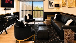 Un salon comprenant un canapé en cuir noir, un fauteuil noir, un pouf noir, une table basse noire, un téléviseur noir et une grande fenêtre donnant sur la forêt.
