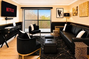 A living room with a black leather sofa, a black armchair, a black ottoman, a black coffee table, a black TV, and a large window with a view of trees.