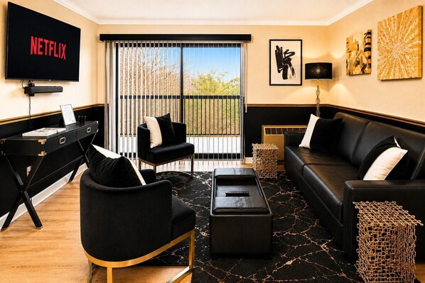 Un salón con un sofá de cuero negro, un sillón negro, un reposapiés negro, una mesa de centro negra, un televisor negro y un ventanal con vistas a árboles.