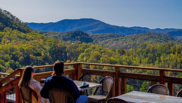 Una terraza de madera con mesa y sillas con vista a una cadena montañosa boscosa.
