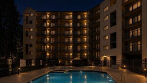 Un hotel de varios pisos con piscina, iluminación exterior y un cartel que indica aguas poco profundas.