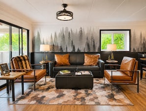 A living room with a black leather sofa, two brown leather chairs, a coffee table, and a hexagonal patterned rug. There are two lamps on side tables, a framed picture on the coffee table, and a window with a view of trees.