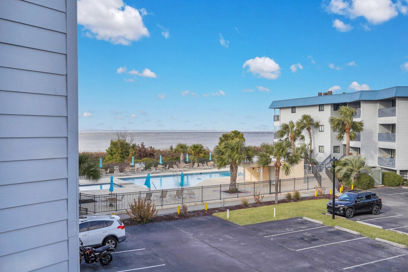 Vista di una piscina con lettini e ombrelloni, un parcheggio e un edificio con balconi affacciati sull'acqua.