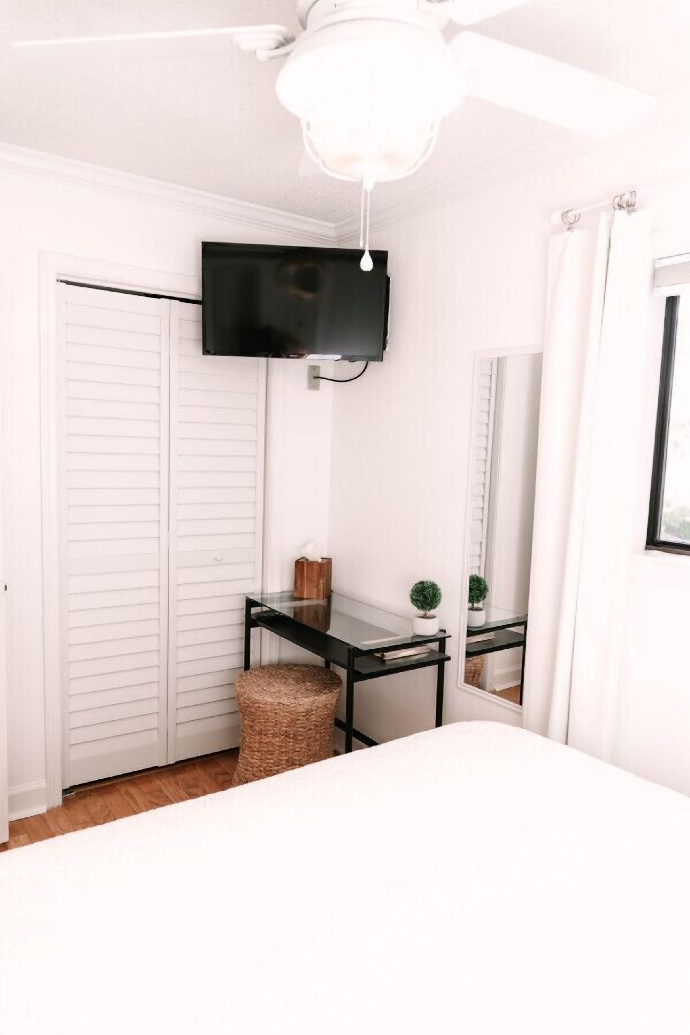 A bedroom with a bed, a desk with a flat-screen TV, a ceiling fan, and a white wardrobe.