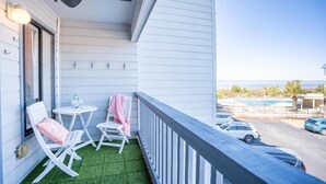 Un balcon avec deux chaises blanches, une petite table et une bouteille d’eau. Le balcon offre une vue sur un parking et une piscine.