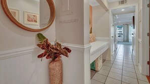 Un couloir avec un mur blanc, un miroir, un vase contenant des fleurs séchées et un sol carrelé.