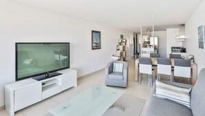 Ein modernes Wohnzimmer mit Flachbildfernseher, einem Glastisch, einem grauen Sofa und einem Essbereich mit Tisch und Stühlen.