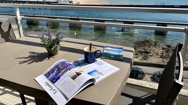 Ein Tisch mit einer Zeitschrift, einer Kaffeetasse und einem Blumentopf auf einem Balkon mit Blick auf Strand und Stadtlandschaft.