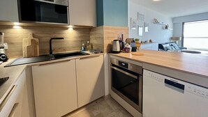 A modern kitchen with a microwave, oven, and dishwasher. There is a sink with a faucet, and a wooden countertop. The kitchen has light blue walls and a window. - Accès direct à la Plage + Garage fermé (Les Sables-d'Olonne)