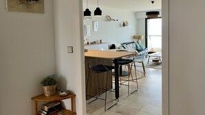 A modern kitchen with a wooden table and bar stools, a dining area with a sofa, and a view of the sea through a large window. - Accès direct à la Plage + Garage fermé (Les Sables-d'Olonne)