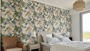 A bedroom with a bed, nightstands, a wicker chair, and a tropical leaf-patterned wallpaper. - Accès direct à la Plage + Garage fermé (Les Sables-d'Olonne)