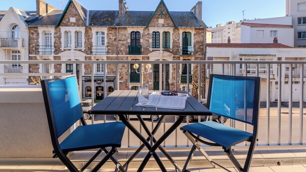 Un balcón con una mesa y dos sillas azules, con vista a una hilera de edificios de piedra con contraventanas verdes y un cielo azul despejado.