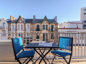 Un balcón con una mesa y dos sillas azules, con vista a una hilera de edificios de piedra con contraventanas verdes y un cielo azul despejado.