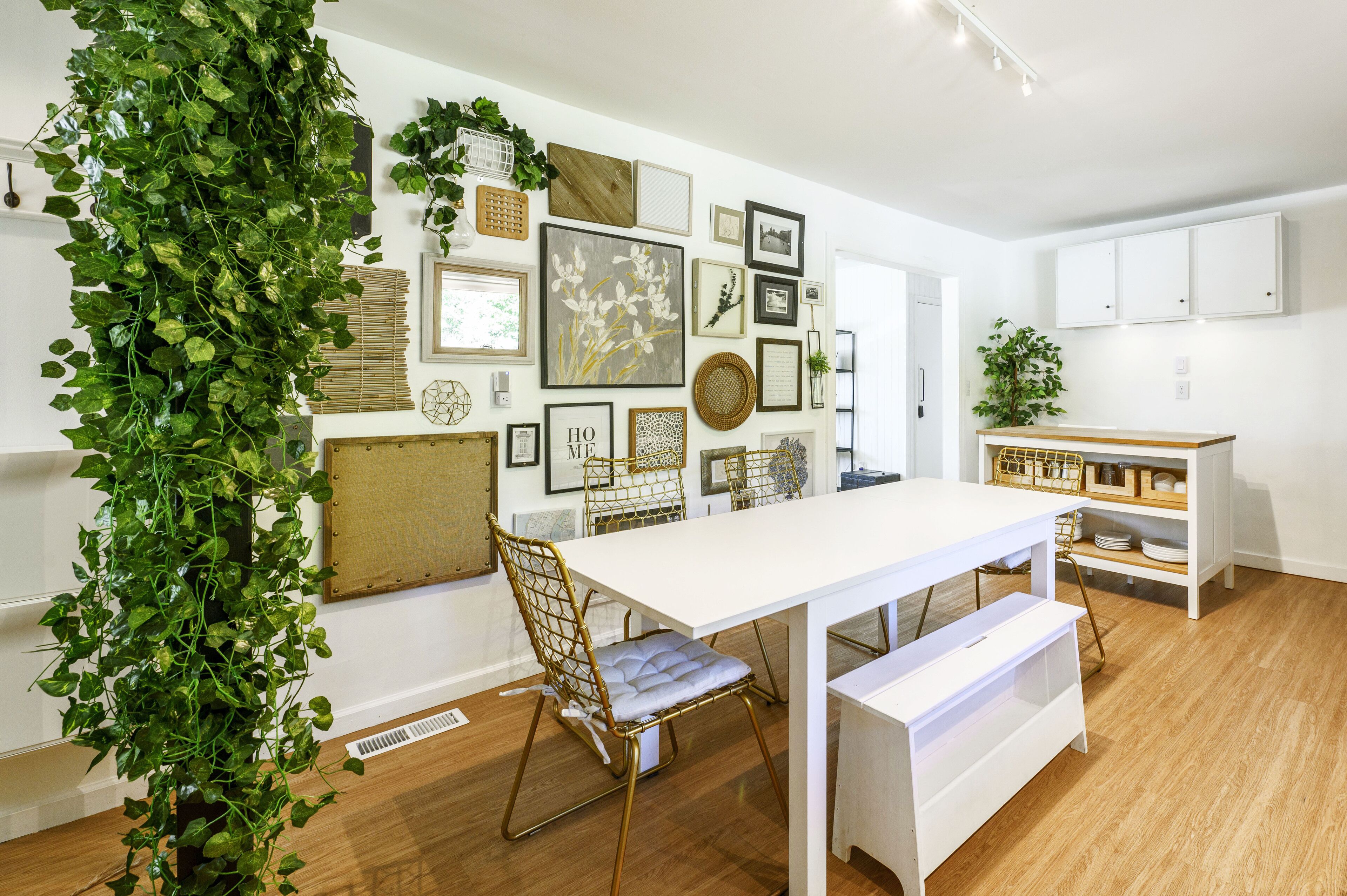 Un comedor con una mesa blanca, una silla con asiento acolchado y un banco. Hay una pared con varios cuadros enmarcados y una planta verde grande.