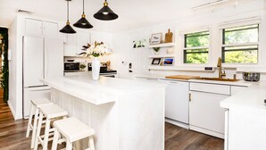 Een moderne keuken met witte kasten, een centraal eiland met barstoelen, een inbouwoven en een raam met uitzicht op groen.