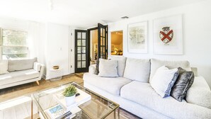 Een moderne woonkamer met een witte bank, een glazen salontafel en een boekenkast. Er hangen ingelijste foto's aan de muur en een deur die leidt naar een andere kamer.