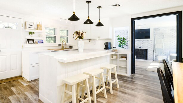 Een moderne keuken met witte kasten, een centraal eiland met krukken, hangende lampen en uitzicht op een buitenruimte.