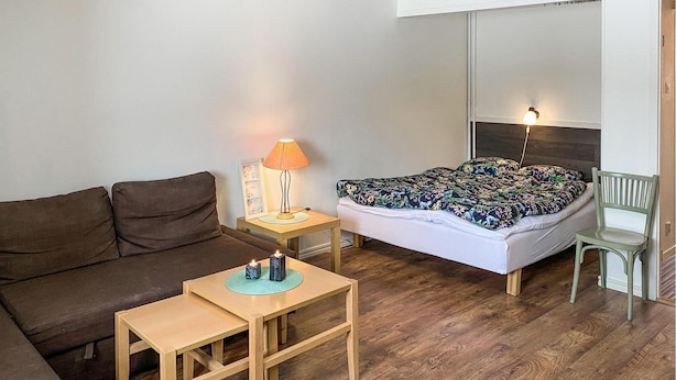 A small, cozy room with a bed, a wooden nightstand with a lamp, a small table, a green chair, and a brown sofa.