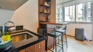 A modern kitchen with a black countertop, stainless steel sink, and wooden shelves. There are two bar stools and a small refrigerator. - OBA 142 - Super Estúdio Frei Caneca (São Paulo)