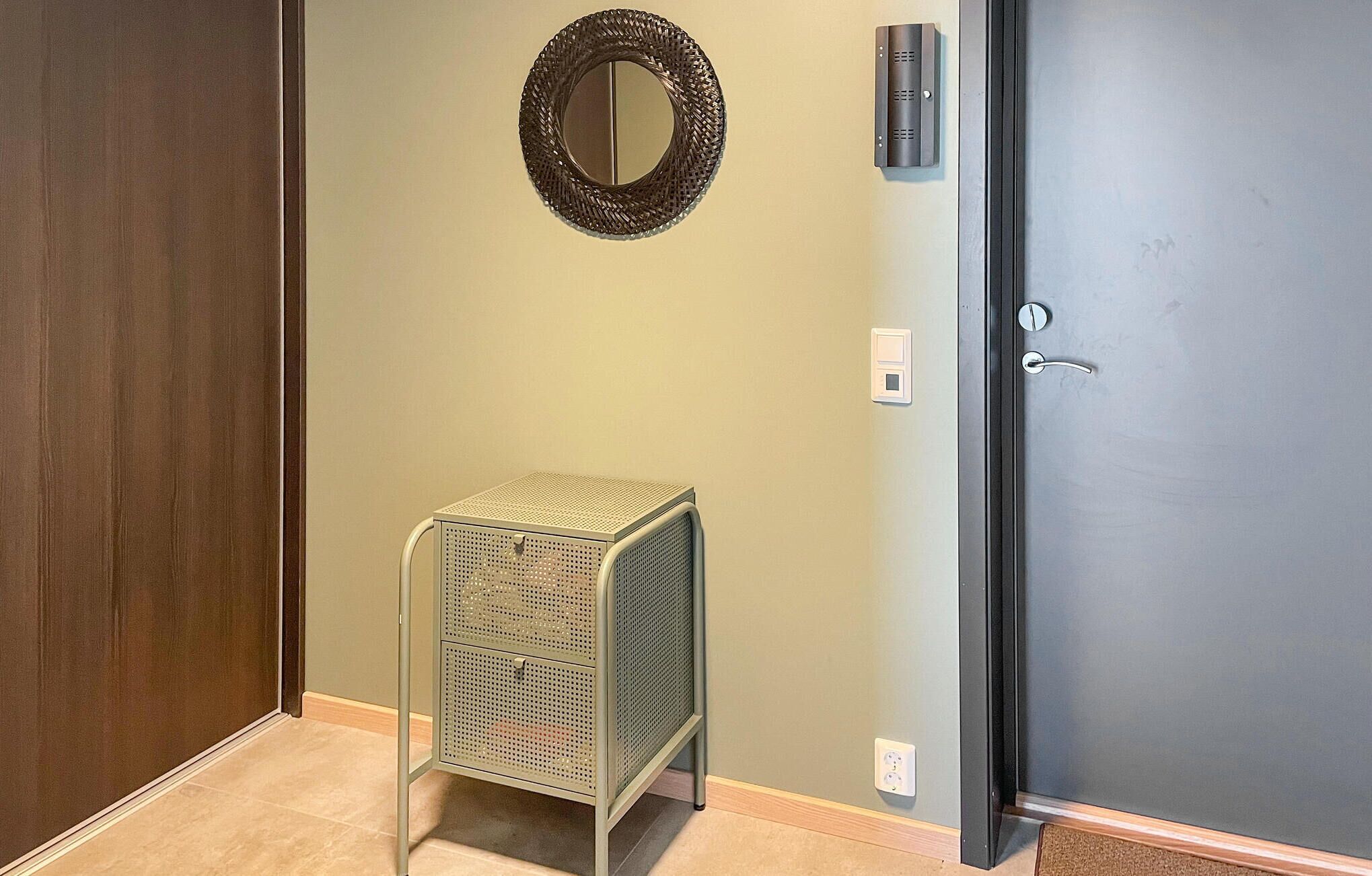A room with a dark door, a small metal storage unit, a wall-mounted light, and a decorative mirror.