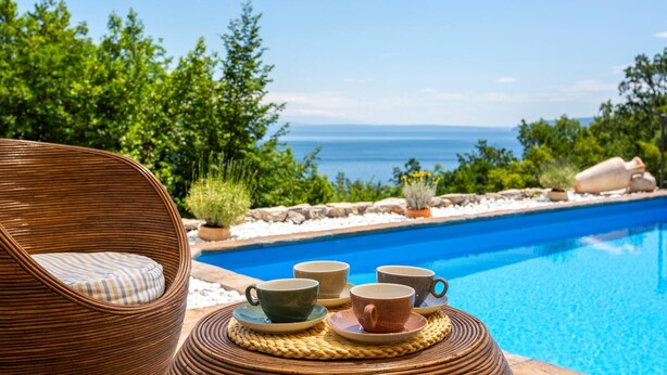A wicker chair with a cushion, a table with four ceramic cups and saucers, and a pool with a view of the sea and greenery. - Amazing home in Lovran (Lovran)