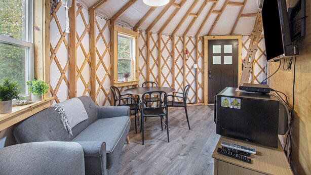 A cozy room with a wooden ceiling, a sofa, a dining table with chairs, a television, and a window with plants. - Luxury Glamping Yurt with Loft, Wood-Fired Hot Tub (Sears)