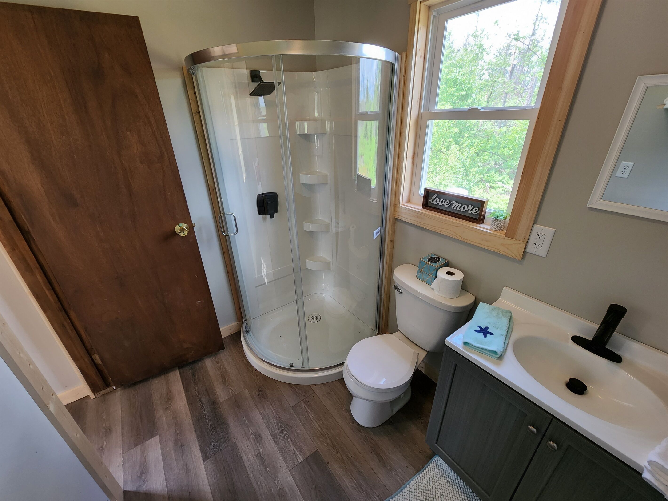 A bathroom with a glass shower enclosure, a toilet, a sink, and a window with a view of greenery.