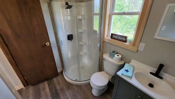 A bathroom with a glass shower enclosure, a toilet, a sink, and a window with a view of greenery.