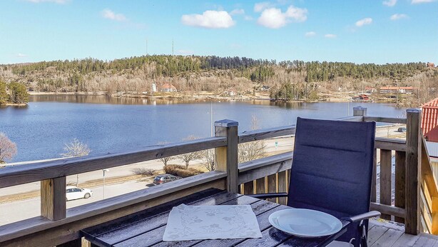 Una terraza de madera con una silla y una mesa, con vistas a un lago con orilla arbolada y edificios a lo lejos.