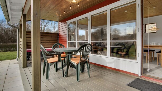 Un patio exterior con terraza de madera, mesa y sillas, y ventanas amplias con vistas al exterior.