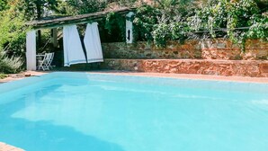 A clear swimming pool with a stone wall and a pergola with white curtains.