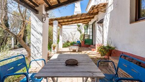 A covered patio with a wooden table and blue chairs, surrounded by potted plants and a white wall.
