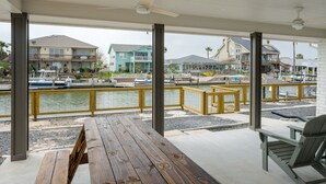 A covered outdoor area with a wooden deck, a table, and a chair, overlooking a marina with boats and houses in the background. - NEWLY REMODELED - Rockport Waterfront Oasis (Rockport)