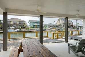 Un espacio al aire libre techado con terraza de madera, una mesa y una silla, con vista a un puerto deportivo con barcos y casas al fondo.
