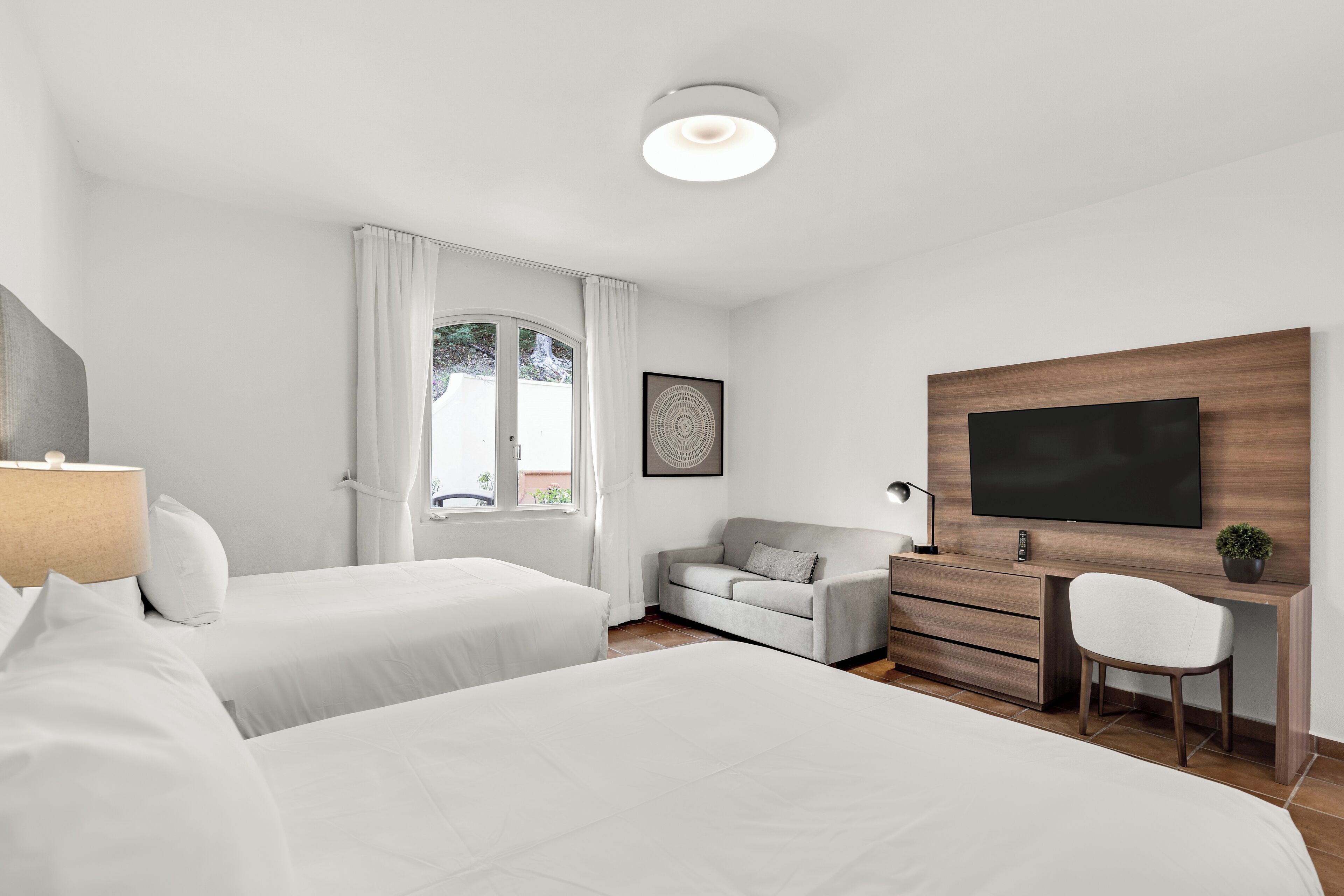 A modern hotel room with two single beds, a sofa, a desk with a chair, a flat-screen TV, and a window with curtains.
